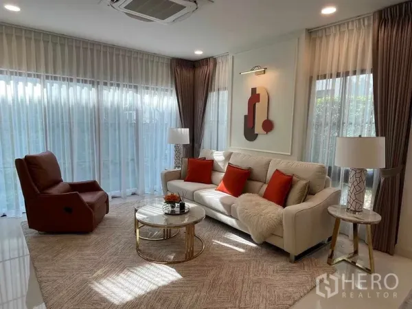 Detached House for rent in Saphan Sung, Bangkok - Sunlit living room with cream sofa, orange cushions, recliner and round coffee table by floor-to-ceiling windows.