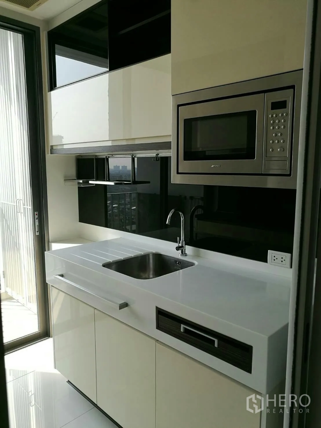 Condominium for rent in Phra Khanong, Bangkok - Bright kitchen area with white countertop, sink, built-in microwave and sliding door opening to a balcony.