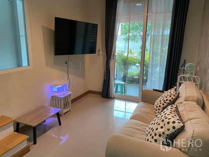 Townhouse for sale in Saphan Sung, Bangkok - Cozy TV corner with sofa and sliding glass doors opening to a small patio with plants.