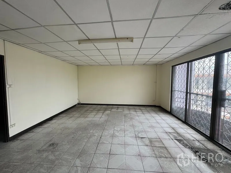 Shophouse for rent in Watthana, Bangkok - Large open office-style room with grid ceiling, tiled floor and windows with security grilles.