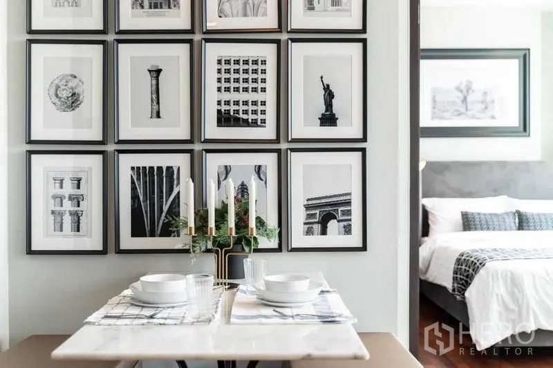 Condominium for rent in Ratchathewi, Bangkok - Dining table set for two beneath a gallery of black-and-white art, leading toward the bedroom.