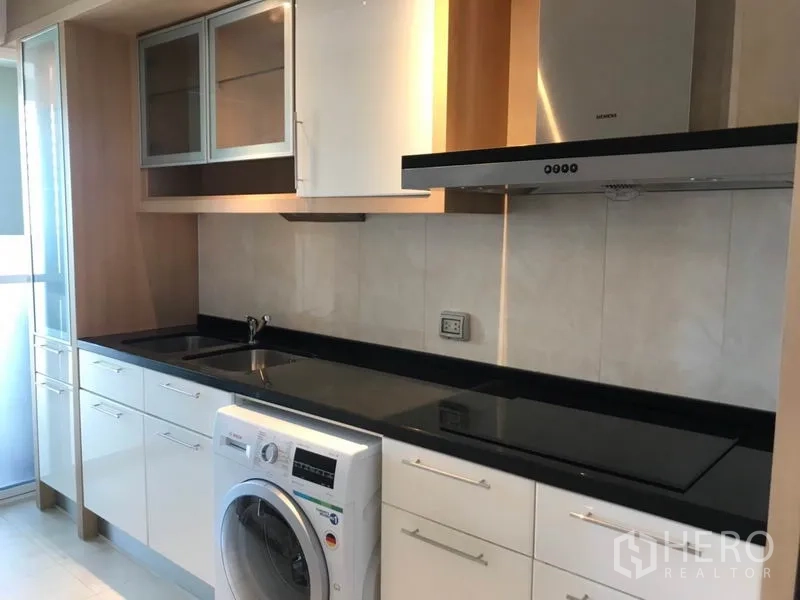 Condominium for rent in Khlong Toei, Bangkok - Modern kitchen with black granite countertop, cooktop, range hood and front-loading washing machine.