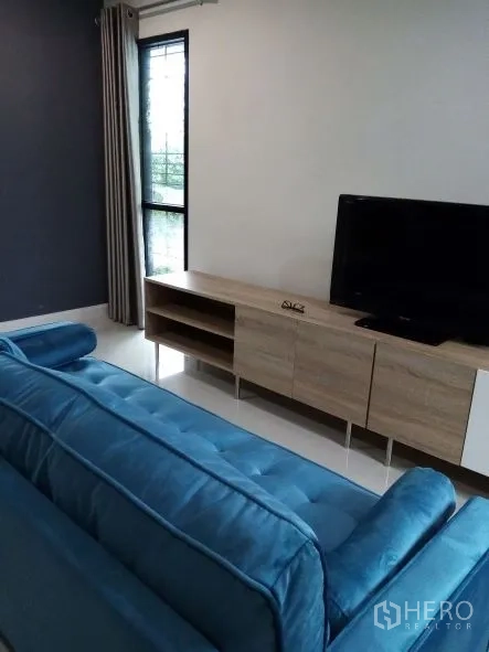 บ้าน ให้เช่าในChok Chai, Nakhon Ratchasima - ห้องนั่งเล่นมีโซฟาสีฟ้า ตู้วางทีวีไม้ และหน้าต่างบานใหญ่พร้อมผ้าม่าน