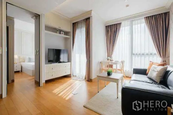 Condominium for rent in Khlong Toei, Bangkok - Living room with TV console, dining nook by tall windows, and doorway to the bedroom.