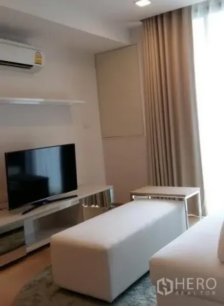 Condominium for rent in Watthana, Bangkok - Bright living room with TV console, white ottoman, and floor-to-ceiling curtains.