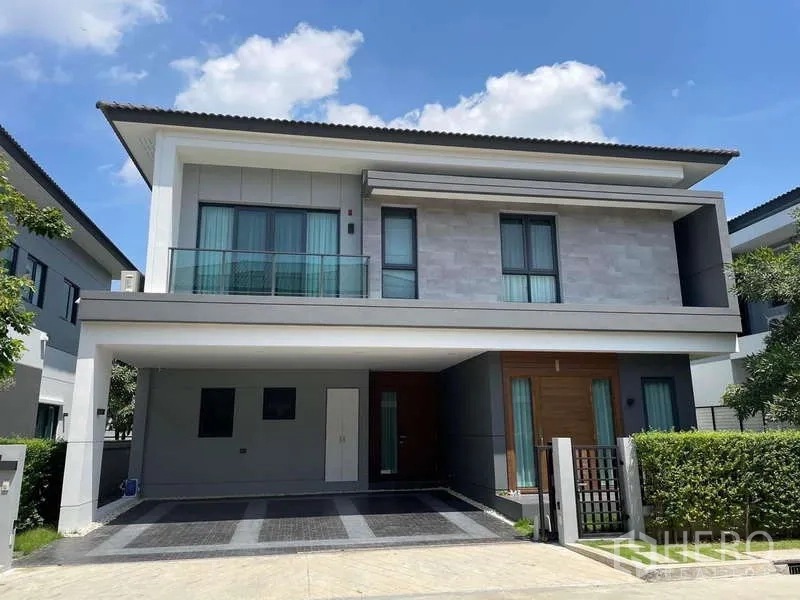 Detached House for rent in Bang Phli, Samut Prakan - Modern two-storey detached house in Bang Phli with balcony, hedge garden and covered carport under blue sky.