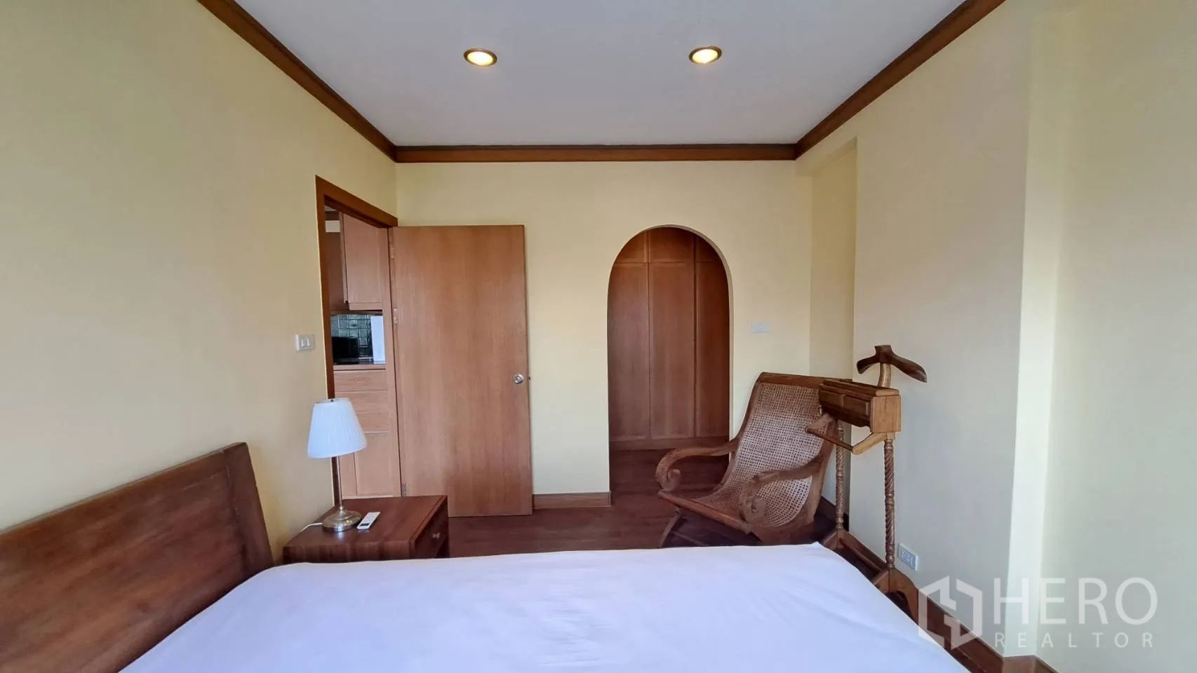 Condominium for rent in Khlong Toei, Bangkok - Cozy bedroom with recessed lights, wooden door, and rattan lounge chair.