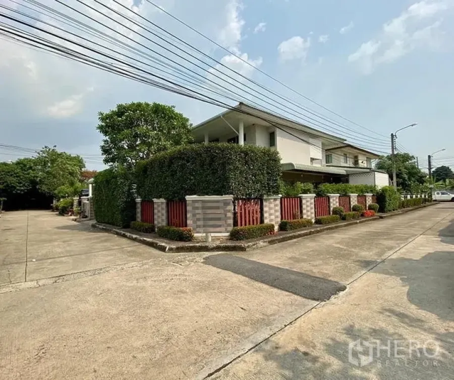 Detached House for sale in Phasi Charoen, Bangkok - Corner‑lot view of the property with privacy hedge, brick fence, and wide concrete road in a quiet neighborhood.