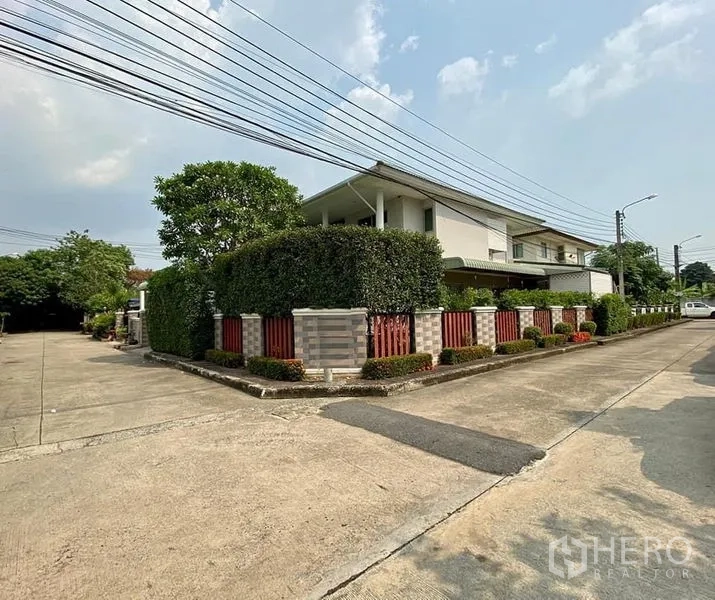 Detached House for sale in Phasi Charoen, Bangkok - Corner‑lot view of the property with privacy hedge, brick fence, and wide concrete road in a quiet neighborhood.