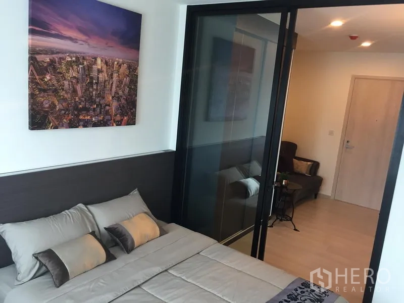 Condominium for rent in Huai Khwang, Bangkok - Bedroom with double bed, cityscape artwork and glass sliding partition opening to the living room.