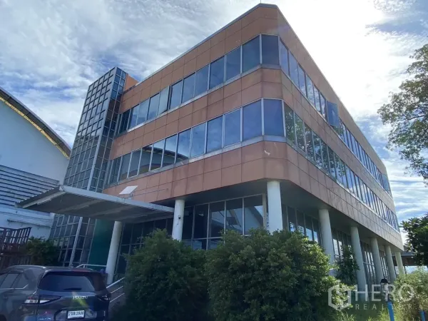 工厂 出售在Samut Prakan - 位于Phraeksa工厂园区的三层玻璃幕墙办公楼，入口设雨棚与停车位，环绕绿化与蓝天。