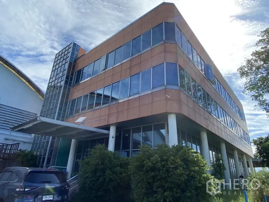 工厂 出售在Samut Prakan - 位于Phraeksa工厂园区的三层玻璃幕墙办公楼，入口设雨棚与停车位，环绕绿化与蓝天。