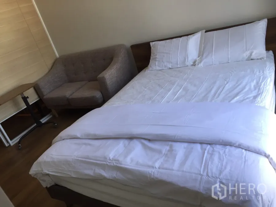 Condominium for rent or sale in Khlong Toei, Bangkok - Cozy Khlong Toei condo bedroom with white bedding, gray sofa and rolling side table on wood floors.