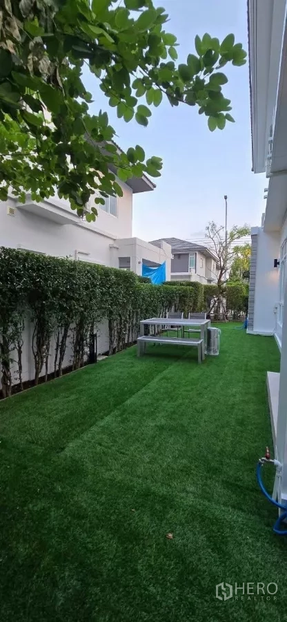 Detached House for rent in Samut Prakan - Long green lawn between houses with hedges and an outdoor bench.
