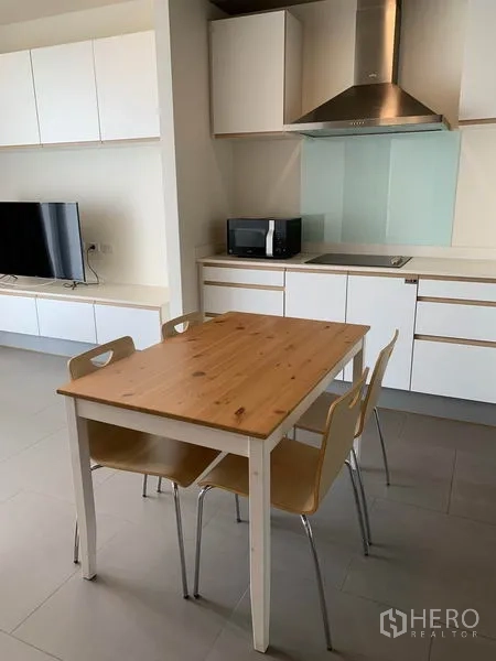 Condominium for rent in Watthana, Bangkok - Dining space with wooden table and four chairs beside a minimalist white kitchen and TV.