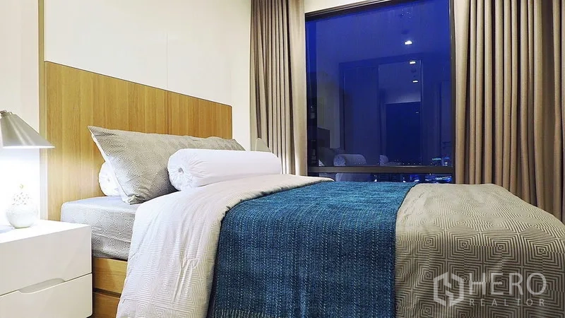 คอนโด ให้เช่าหรือขายในKhlong Toei, Bangkok - ห้องนอนโทนอบอุ่น เตียงผ้าคลุมสีน้ำเงิน โต๊ะข้างเตียง และหน้าต่างบานใหญ่เห็นวิวเมืองยามค่ำคืน