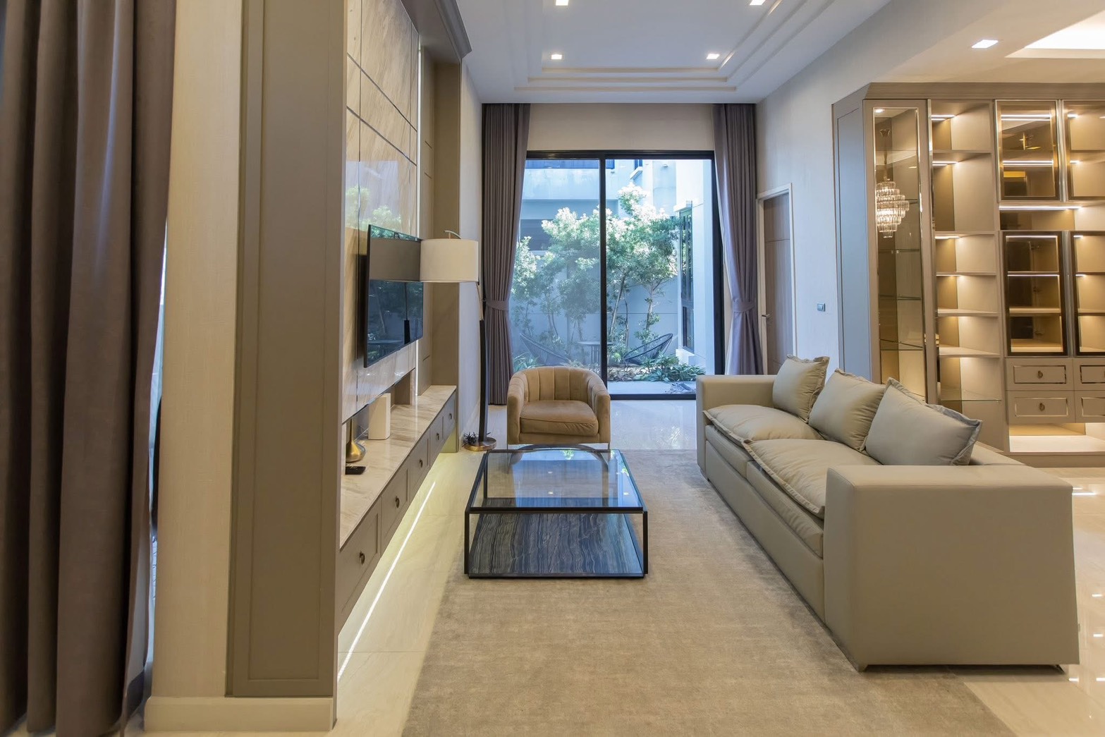 独立式住宅 出租在Lat Phrao, Bangkok - 明亮客厅，长沙发、电视墙、玻璃茶几，推拉门通向小花园。