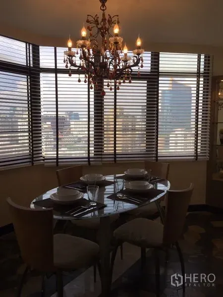 Condominium for rent in Watthana, Bangkok - Dining nook with chandelier, round table set for four and city views through wide windows with blinds.