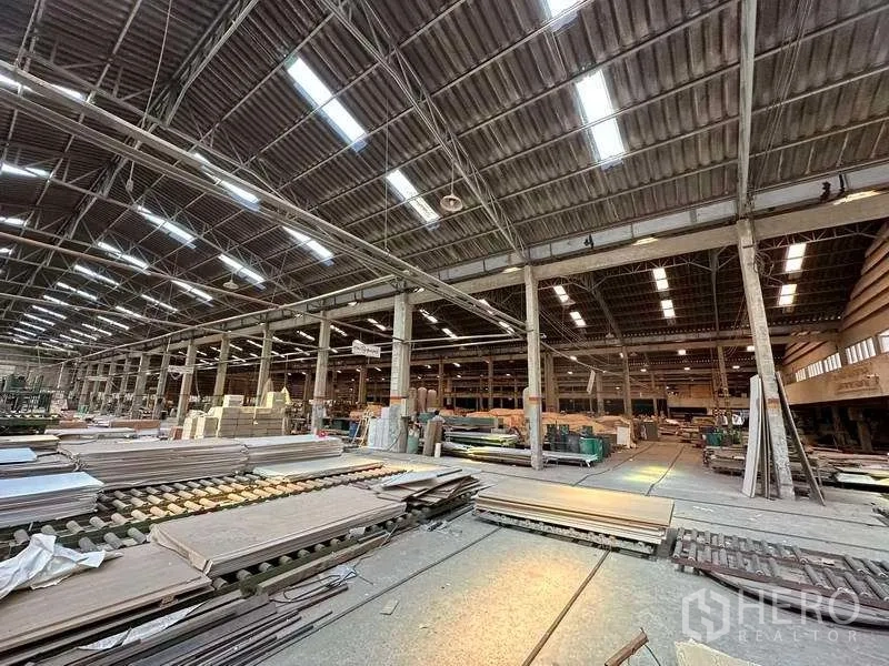 工厂 出租在Sai Noi, Nonthaburi - 生产区域堆料与设备，灯光明亮的钢桁架屋顶下。