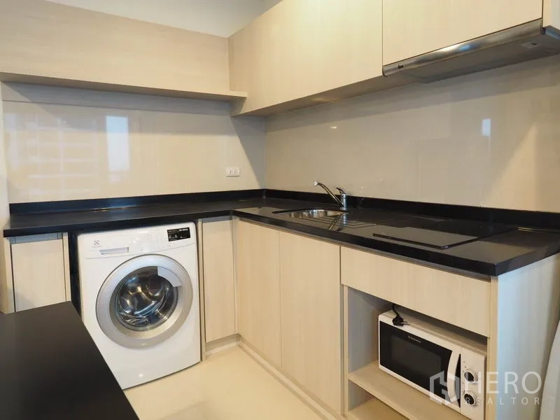 Condominium for rent in Sathon, Bangkok - L-shaped kitchen with light wood cabinetry, black countertops, cooktop, hood, washer and microwave.