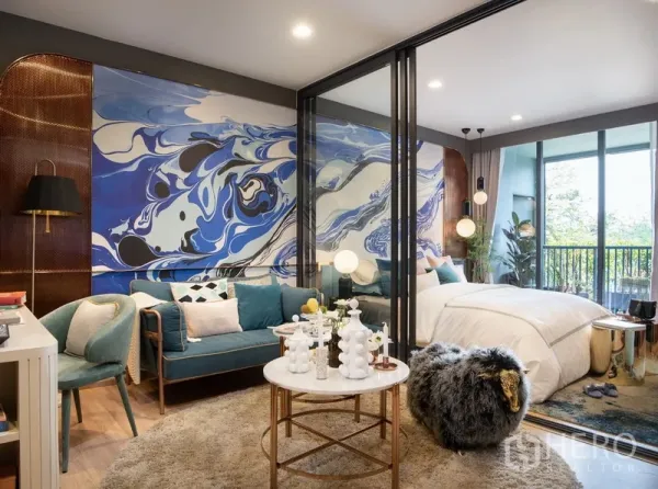 公寓 出售在Ratchathewi, Bangkok - 客厅与卧室以玻璃分隔，蓝色抽象背景墙，带观景阳台的绿意视野。
