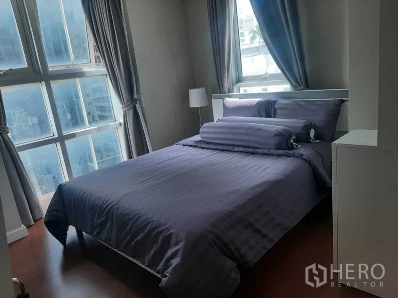 Condominium for rent or sale in Huai Khwang, Bangkok - Corner bedroom with large multi-pane windows, grey curtains, and city outlook.