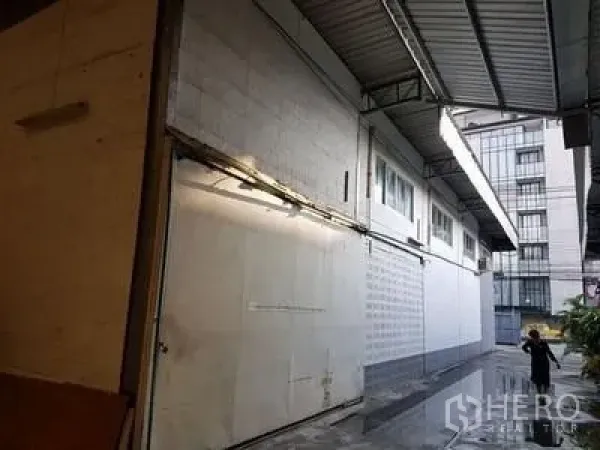Warehouse for rent in Chatuchak, Bangkok - Covered loading alley along a white warehouse wall with high metal roof, drainage channel and neighboring modern building near Ratchadaphisek.