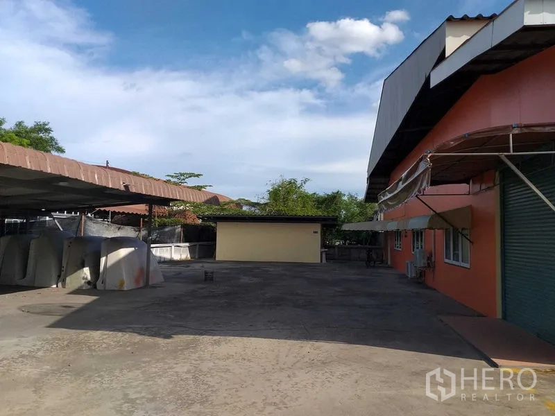 货栈 出租在Khlong Sam Wa, Bangkok - 建筑侧边宽阔硬地院坝，带雨棚与卷帘装卸口，蓝天晴空。