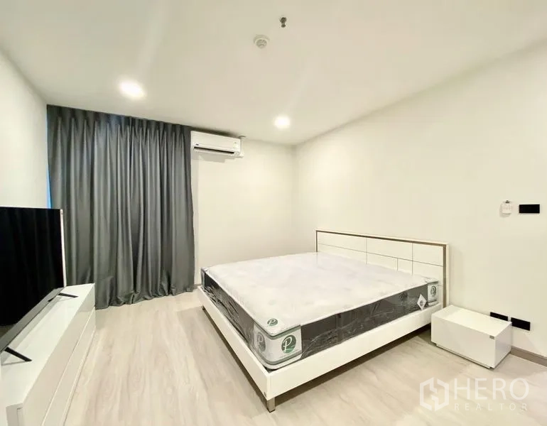 คอนโด ให้เช่า - ห้องนอนหลักพร้อมชั้นวางทีวี โต๊ะหัวเตียง และม่านทึบ