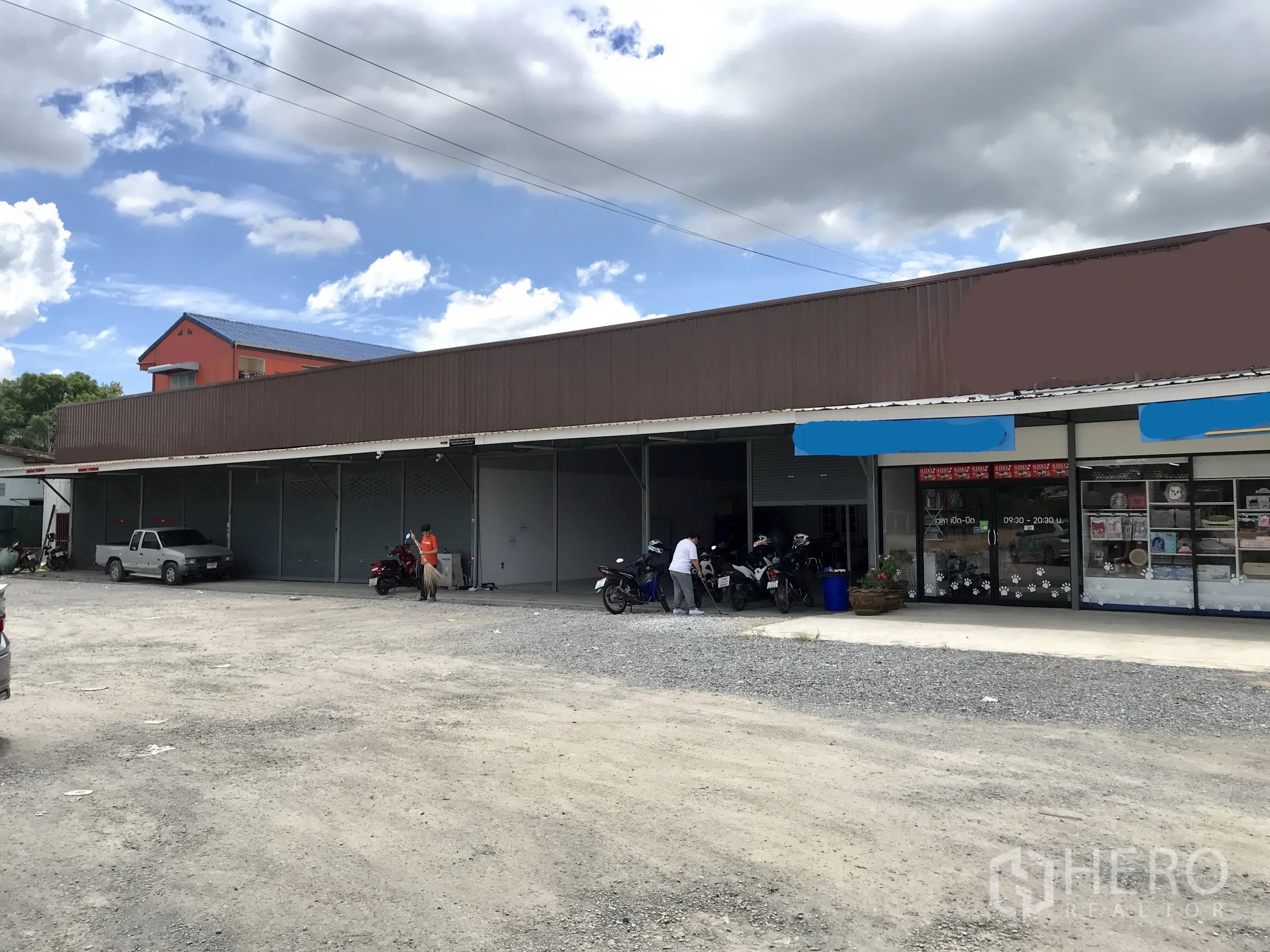 คลังสินค้า ให้เช่าในLam Luk Ka, Pathum Thani - โกดังชั้นเดียวหน้าถนน มีประตูม้วนหลายบานและลานจอดรถโรยหินกว้าง ข้างอาคารร้านค้าเล็ก.