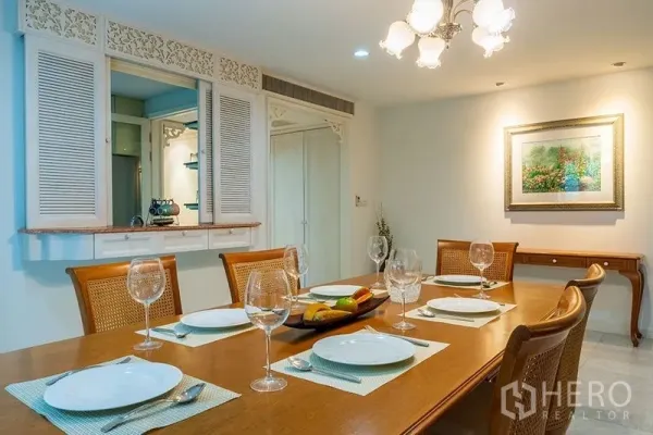Apartment for rent in Bang Rak, Bangkok - Dining room with wooden table set for six, chandelier lighting and kitchen pass‑through with louvered shutters.