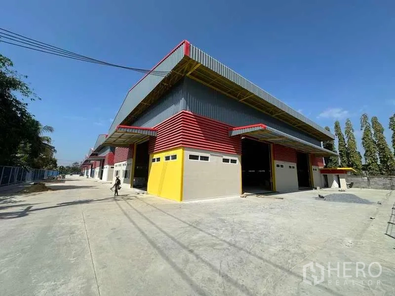 货栈 出租在Lam Luk Ka, Pathum Thani - 仓库转角视角，带雨棚的装卸区，宽大前场与绿树围合边界。