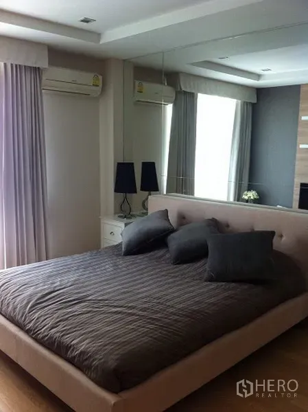 Condominium for rent or sale in Pathum Wan, Bangkok - Bright condo bedroom with upholstered bed, mirrored wall, curtains and air conditioning.