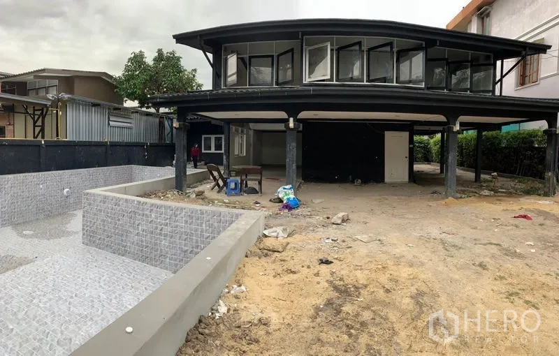 独立式住宅 出售在Suan Luang, Bangkok - 房屋正面近景：上层悬挑、下方覆盖式停车位，旁有空置贴砖泳池与沙土地。