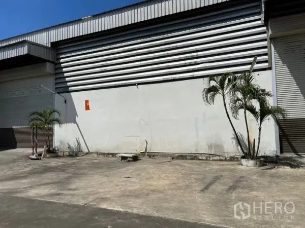 货栈 出租在Prawet, Bangkok - 仓库外立面，条纹金属板墙体与卷闸门，前方大面积硬化地面并点缀棕榈