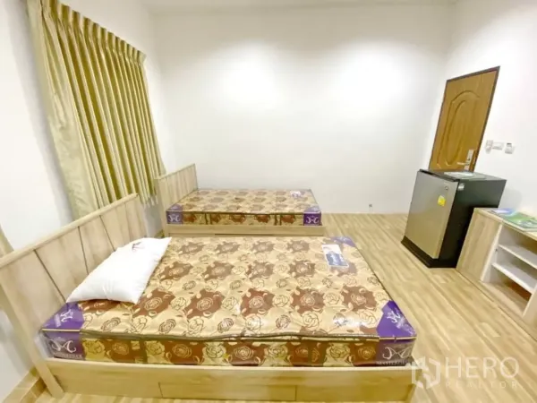 Apartment for sale in Khlong Toei, Bangkok - Twin beds facing a wooden door with a mini fridge and shelving in a furnished room.