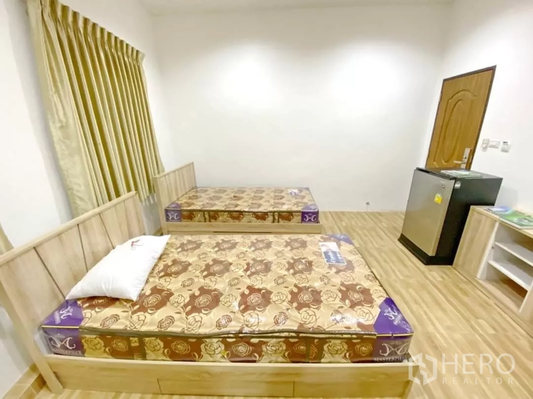 Apartment for sale in Khlong Toei, Bangkok - Twin beds facing a wooden door with a mini fridge and shelving in a furnished room.