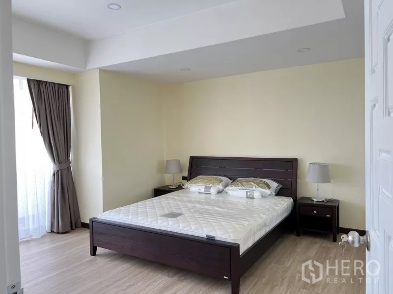 คอนโด ให้เช่าในKhlong Toei, Bangkok - ห้องนอนใหญ่ เตียงไม้เข้ม โต๊ะข้างเตียงพร้อมโคมไฟ และผ้าม่านยาวจรดพื้น