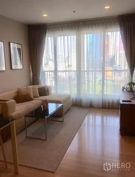Condominium for rent in Sathon, Bangkok - Living room with floor-to-ceiling windows, sheer curtains and city skyline view beside a sofa and TV console.