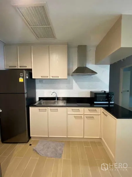Condominium for rent in Khlong Toei, Bangkok - L-shaped kitchen with black stone countertop, range hood, upper and lower cabinets and fridge.
