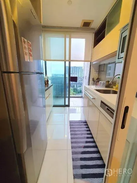Condominium for sale in Bangkok - Compact white kitchen with built-in cabinets, stainless fridge, and a sliding glass door opening to a city-view balcony.