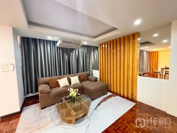 Condominium for rent in Huai Khwang, Bangkok - Bright living room with brown L-shaped sofa, wooden slat divider, recessed ceiling lights, parquet flooring and a round glass coffee table.