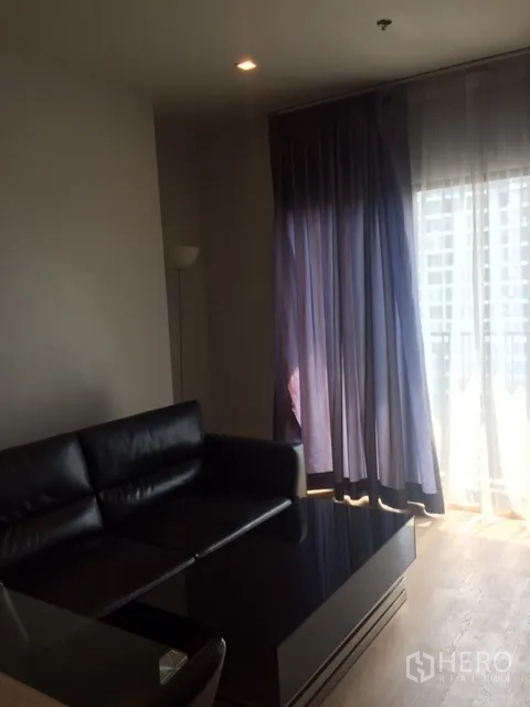 คอนโด ให้เช่าในBangkok - มุมห้องนั่งเล่น โซฟาหนังทรงแอล โต๊ะกลาง และผ้าม่านทึบข้างประตูกระจกบานเลื่อน