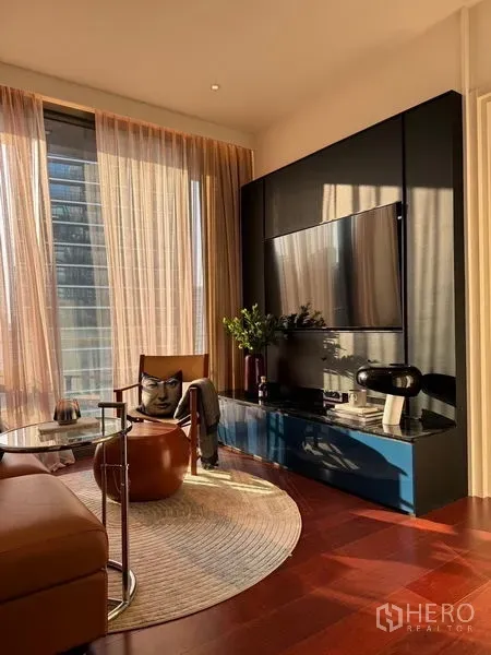 Condominium for sale in Watthana, Bangkok - Sunlit living room with floor-to-ceiling windows, sheer curtains, wall-mounted TV on a black panel, blue console, leather chair and round rug in a Thonglor condo.