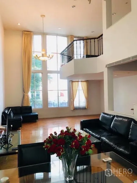 Townhouse for rent in Khlong Toei, Bangkok - Double-height lounge with chandelier, floor-to-ceiling windows and mezzanine railing.