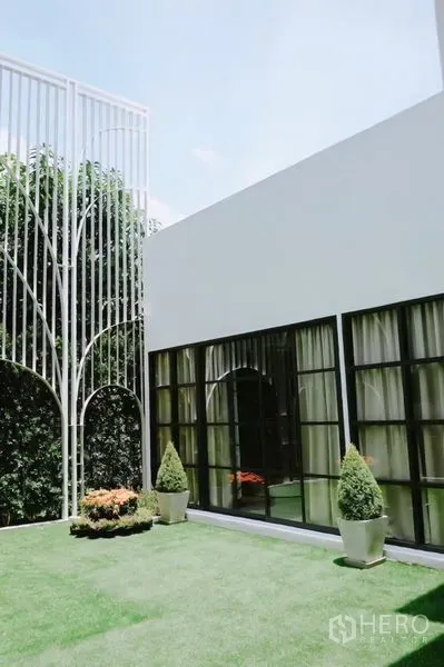Detached House for rent in Huai Khwang, Bangkok - Modern white detached house with black-framed windows opening to a green lawn and decorative metal trellis.