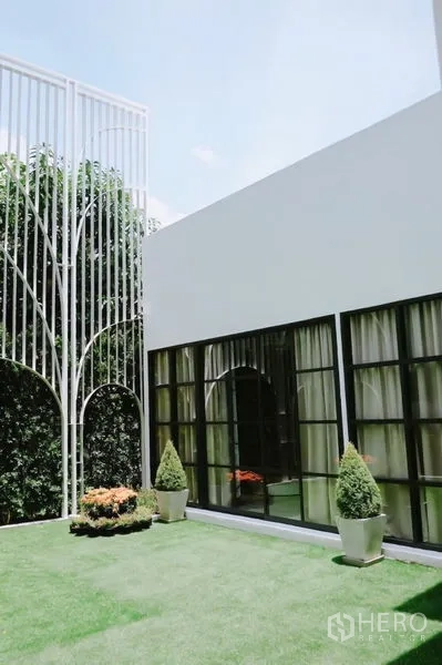 Detached House for rent in Huai Khwang, Bangkok - Modern white detached house with black-framed windows opening to a green lawn and decorative metal trellis.