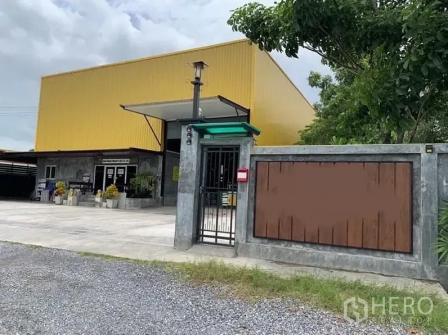 โรงงาน ขายในThanyaburi, Pathum Thani - ประตูรั้วและลานคอนกรีตด้านหน้าโรงงานผนังสีเหลือง พร้อมกำแพงรอบและต้นไม้