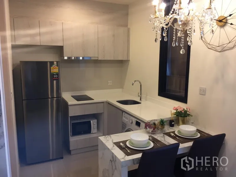 Condominium for rent in Huai Khwang, Bangkok - L-shaped kitchenette with stone countertop, sink, electric hob, fridge, microwave and washer beside a two-seat breakfast bar and chandelier.