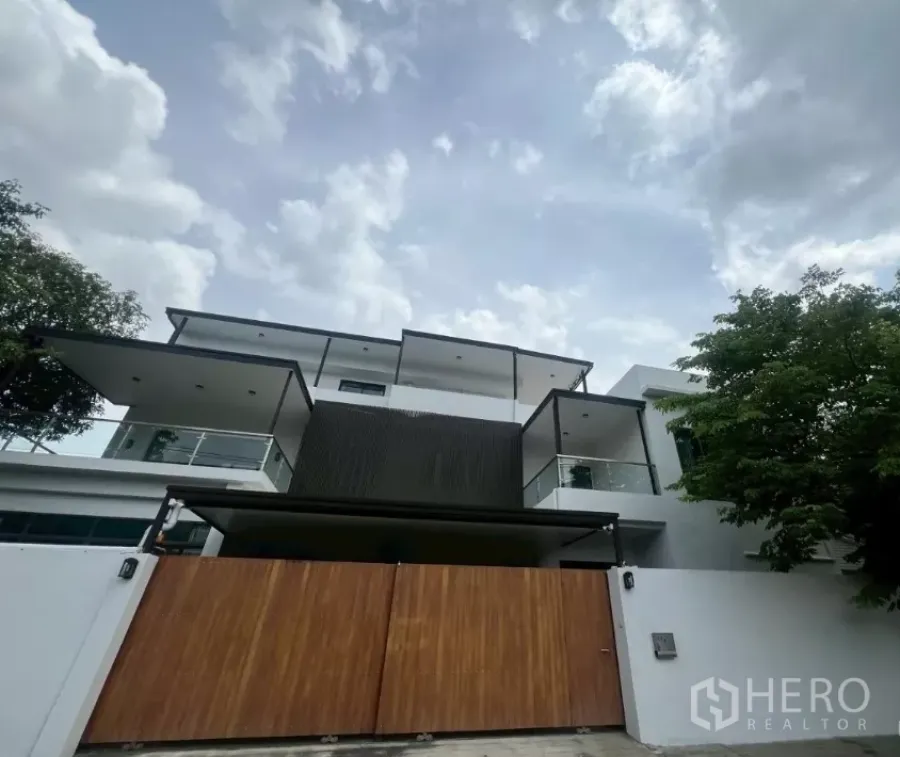 Home Office for rent in Watthana, Bangkok - Modern multi‑level home office exterior with white walls, glass balconies and a wide wooden gate under a cloudy sky.